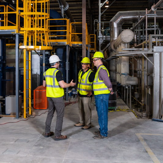 Three men in yellow safety gear are standing on the manufacturing floor