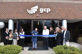 Group of people on holding a blue ribbon on each end and a man using scissors to cut he ribbon in the middle while in front of a brick office building