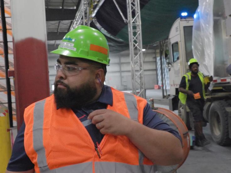 A man with a yellow hard hat and orange vest is on manufacturing plant floor