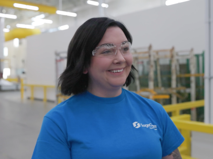 A woman wearing a blue SageGlass shirt and safety goggles walks through a glass factory