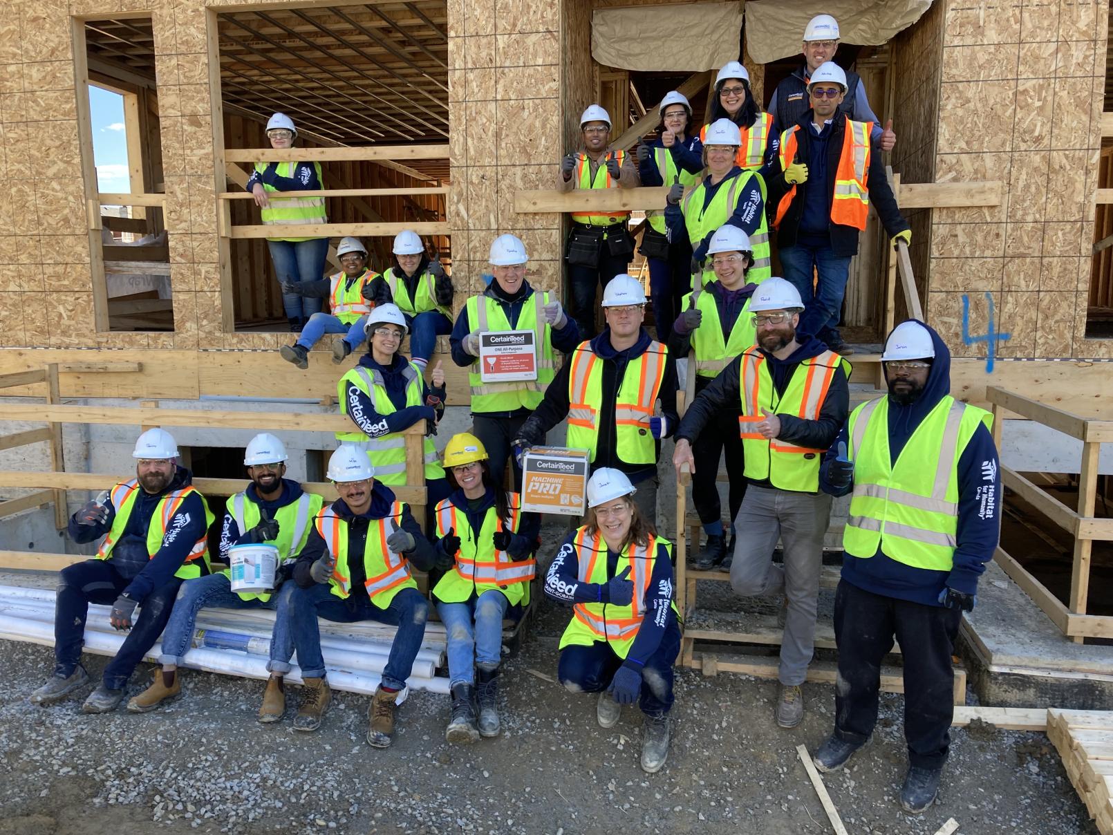 A large group of people wearing safety gear pose for a picture around a home being constructed