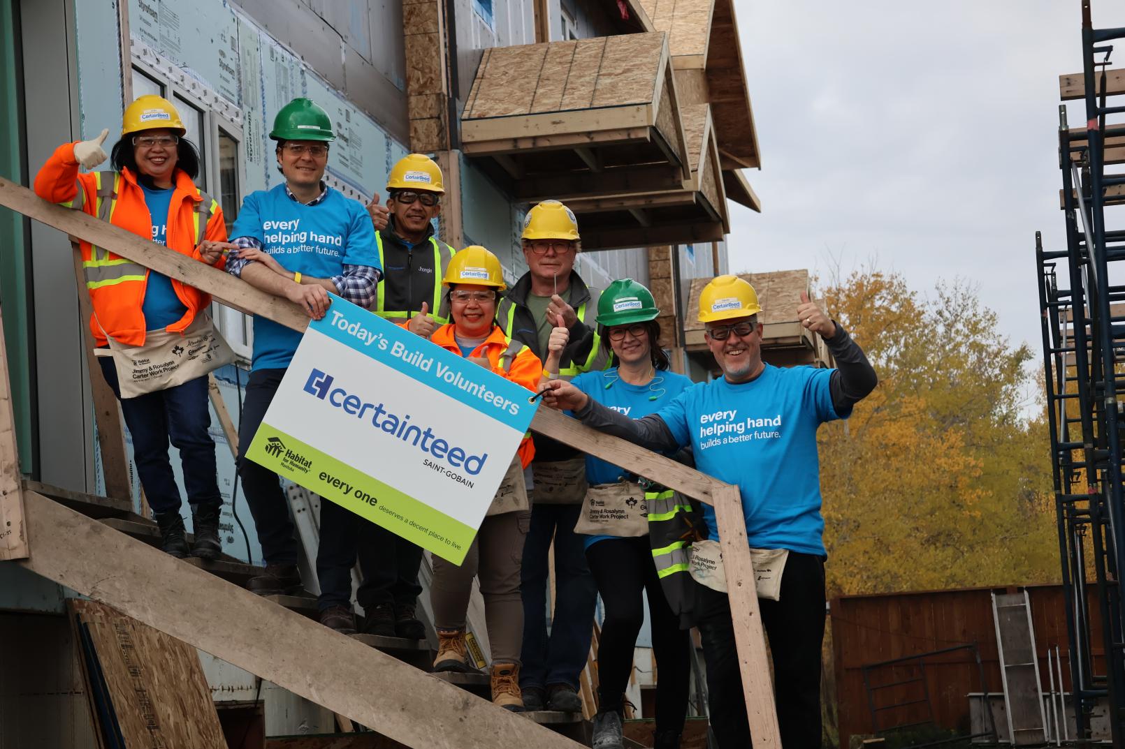 Seven people wearing hard hats are standing on stairs being build holding a sign that says "Today's Build Volunteers" with CertainTeed's logo