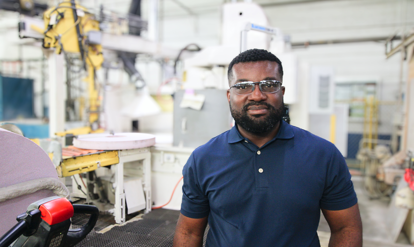 A Black man wearing a blue polo shirt stands in a factory setting in front of white machinery
