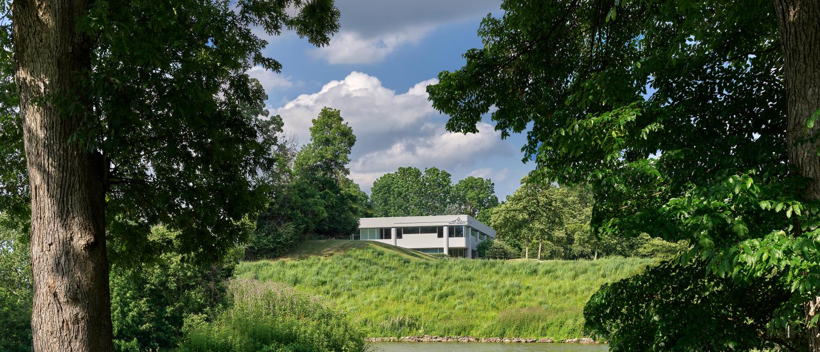 A white building with the Saint-Gobain logo sits behind a hill.  Trees and a small pond are in the foreground. 